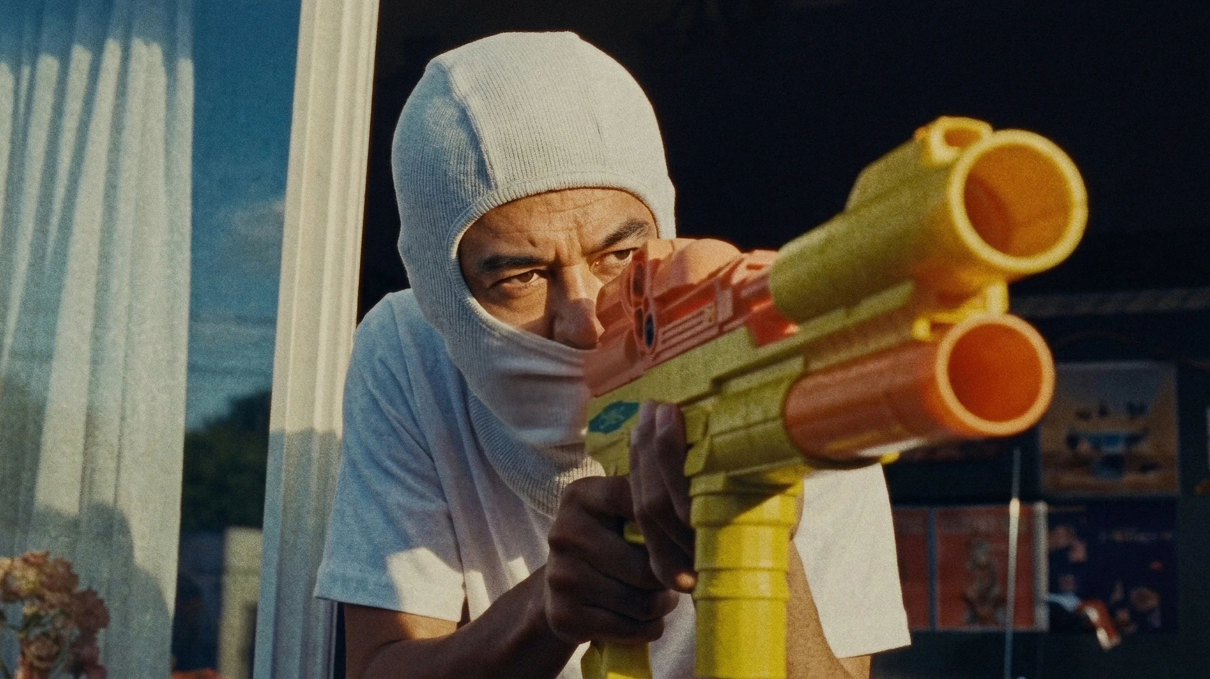 Keiti in a white balaclava aiming a yellow and orange toy water gun out of a window